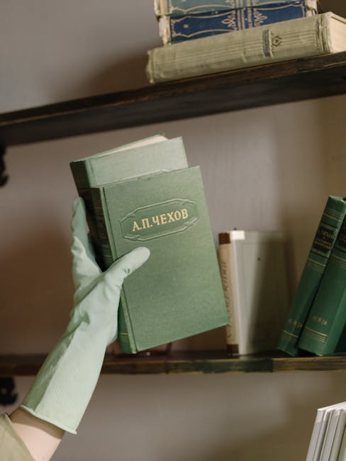 A person wearing light green rubber gloves and a beige sleeve is holding a stack of white paper documents or menus, positioned near a white wall or partition. The surface appears clean and smooth, with soft ambient lighting illuminating the area. In the background, a spray bottle and cleaning supplies are visible on a shelf, indicating a cleaning environment. The setting suggests a moment of document handling or organisation within a cleaning or maintenance context, consistent with professional domestic cleaning services provided by Cleaner Harringay, emphasizing surface cleaning and hygiene practices.