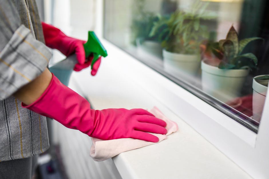A person wearing pink rubber gloves is cleaning a white windowsill with a soft cloth, using a green spray bottle for surface cleaning. The window area includes a row of potted plants with green leaves placed on the sill. The scene is well-lit, with natural light streaming in through the window, revealing a clean and tidy setting. The focus is on the meticulous cleaning process, highlighting the attention to hygiene and surface maintenance, as performed by Cleaner Harringay in domestic and commercial environments.