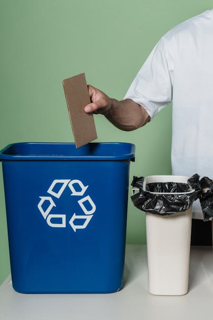 A blue recycling bin positioned beside a white, box-style catering or delivery vehicle with a flat concrete ground surface, in an outdoor setting. The bin appears clean with minor dirt marks, and the