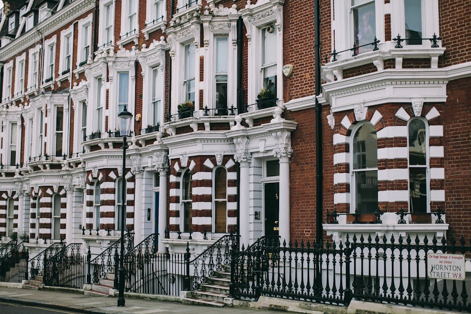 Exterior view of a row of Victorian terraced houses with red brick facades and decorative white stone detailing around the windows and doorways. Each house features large arched windows with white frames and black wrought iron railings along the small front gardens. Black stairs lead up to the front doors, which are painted dark and surrounded by detailed white molding. A traditional street lamp is positioned in front of the buildings, and a street sign indicating 'Hornton Street W8' is visible. The scene is well-lit with natural daylight, emphasizing the clean and well-maintained appearance of the facades, reflecting a high level of surface cleaning and preservation typical of deep cleaning services in historic residential areas, as offered by Cleaner Harringay.