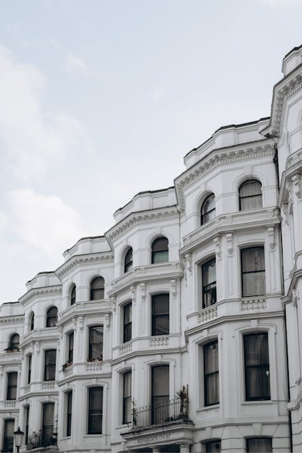 Photograph of a row of Victorian terrace houses with white stucco facades, detailed molding, and arched windows. The buildings are set against a partly cloudy sky, with natural daylight illuminating the exteriors. The image captures the exterior architecture, emphasizing the clean, well-maintained appearance of the facades. This setting highlights the importance of surface cleaning and maintenance for historic residential properties, as part of professional domestic cleaning services offered by Cleaner Harringay, focusing on deep cleaning and sanitisation of architectural surfaces.