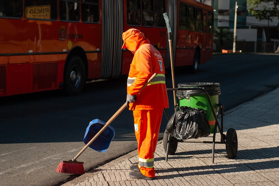 A street worker dressed in bright orange high-visibility clothing, including a hood, is sweeping the sidewalk with a long-handled broom. The worker is positioned next to a wheeled cleaning cart containing a large black plastic bag and several cleaning tools. In the background, a red double-decker bus is visible, and the scene is illuminated by natural daylight, showing a clean and well-maintained urban area. The image emphasizes outdoor cleaning activities, likely part of a sanitation or street maintenance operation, consistent with professional cleaning services offered by Cleaner Harringay in the Green Lanes area.