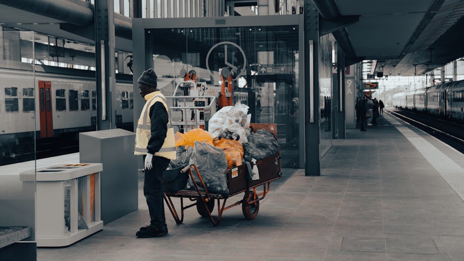 A person dressed in a black hoodie, black pants, gloves, and a high-visibility vest stands on a train station platform next to a large metal trolley loaded with garbage bags and waste materials. The platform features grey tiled flooring, a glass shelter, and several train tracks, with trains visible on both sides and a few additional commuters in the background. The scene is illuminated by natural daylight, and the cleaning activity emphasizes hygiene and maintenance in a public transportation environment, consistent with surface cleaning and sanitisation practices as offered by Cleaner Harringay for residential and commercial spaces in the Green Lanes area, HARRINGAY.