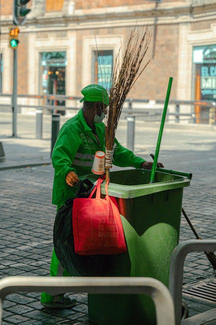 A street worker dressed in bright orange high-visibility clothing, including a hood, is sweeping the sidewalk with a long-handled broom. The worker is positioned next to a wheeled cleaning cart containing a large black plastic bag and several cleaning tools. In the background, a red double-decker bus is visible, and the scene is illuminated by natural daylight, showing a clean and well-maintained urban area. The image emphasizes outdoor cleaning activities, likely part of a sanitation or street maintenance operation, consistent with professional cleaning services offered by Cleaner Harringay in the Green Lanes area.