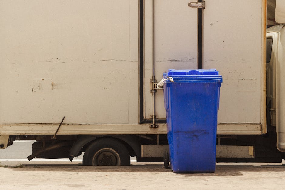 A blue recycling bin is positioned next to a white delivery truck on a paved surface, with the bin's lid closed and slight dirt marks visible on its surface. The truck has a plain white exterior with 