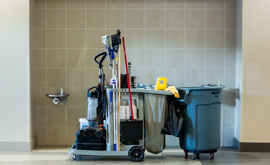 A cleaning trolley positioned in front of a beige tiled wall in a commercial or residential setting, with cleaning tools such as mops, spray bottles, and cloths stored on it. To the left, there is a small stainless steel sink mounted on the wall. To the right, a large blue wheeled trash bin is present. The area is well-lit with neutral lighting, and the surfaces appear clean and tidy, indicating ongoing or recent cleaning activity. The scene reflects professional cleaning service provision, consistent with the services offered by Cleaner Harringay, emphasizing surface cleaning and maintenance.