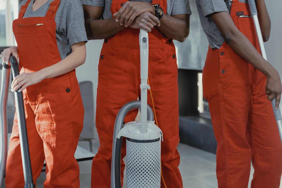 Close-up of three professional cleaners dressed in grey shirts and orange overalls, each holding a vacuum cleaner hose in a brightly lit, modern indoor space with tiled flooring. The central cleaner is gripping a white and grey vacuum unit with a flexible hose, while the other two hold their vacuum attachments. The scene showcases tools used for surface cleaning and deep cleaning, emphasizing hygiene and maintenance. Cleaner Harringay's branding is implied through the professional attire, and the setting appears organized and clean, suitable for residential or commercial cleaning services.